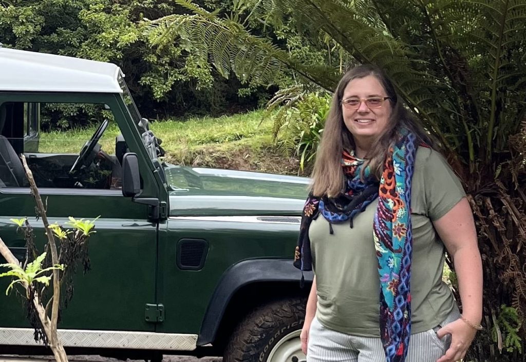 Frau mit buntem Tuch im den Hals vor kleiner Palme und neben einen grünen Jeep